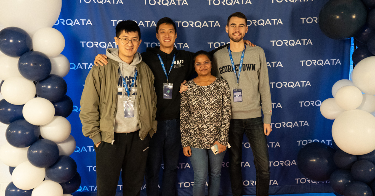 Four students stand together smiling and facing the camera. They are standing in front of a Torqata Hackathon backdrop and framed on either side by columns of blue and white balloons.