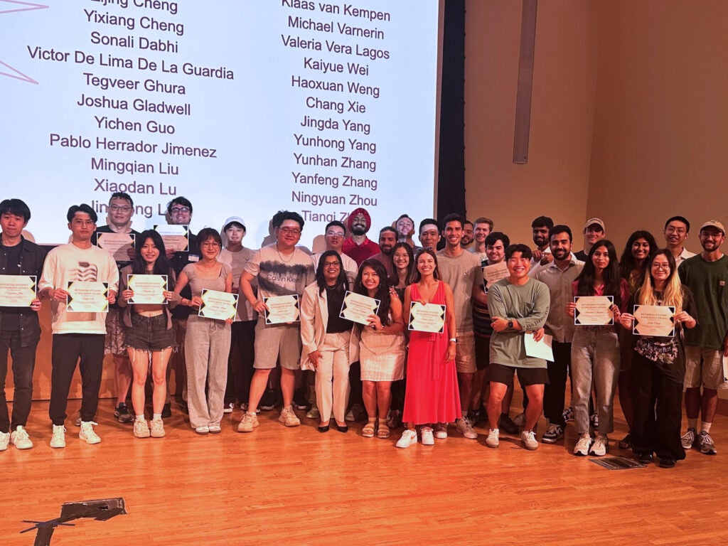 A large group of students and the DSAN director stand smiling in front of a screen listing the names of winners.