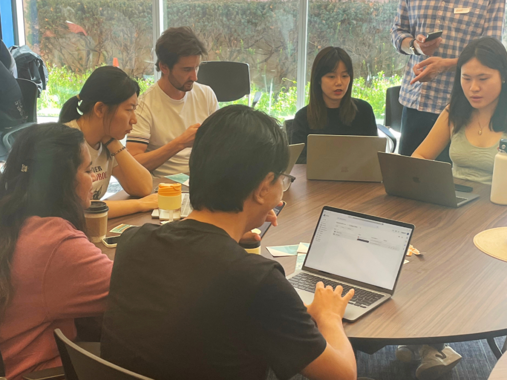 Several people are gathered at a round table in front of a window, working in their laptops at a hackathon.