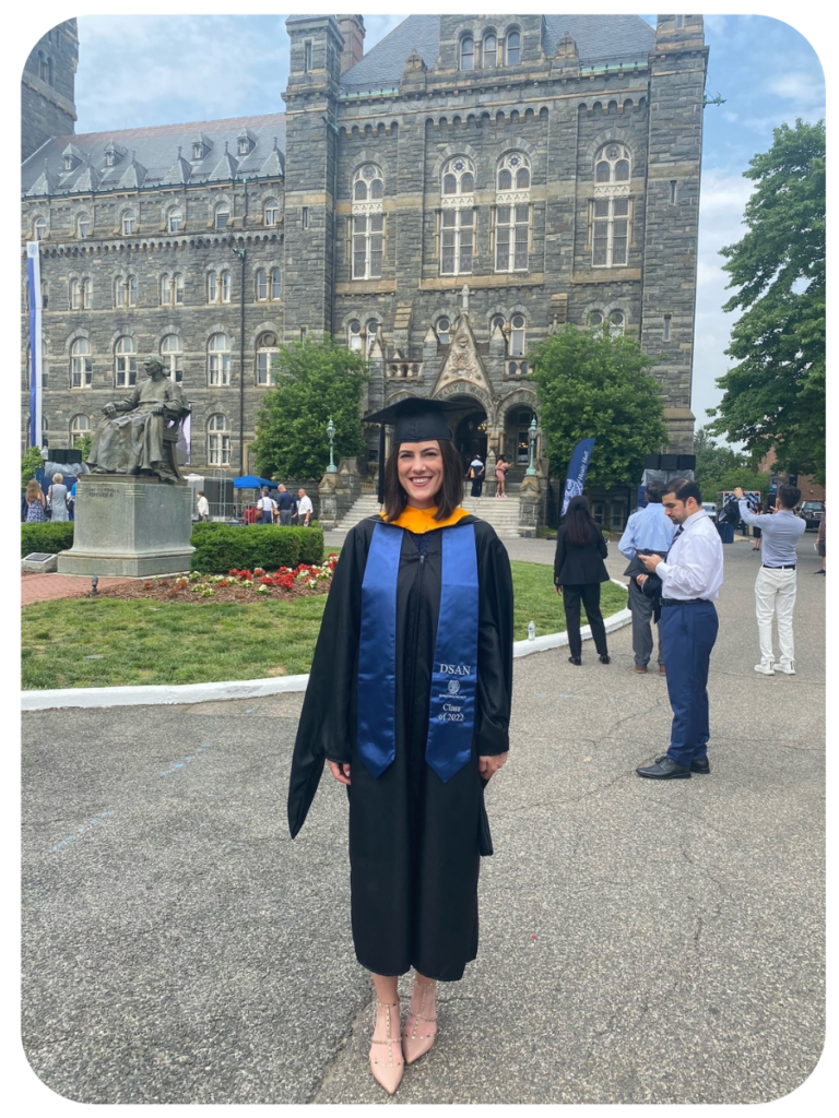 Gabriella stands in a black graduation robe and a bright blue stole. Healy Hall and Healy Lawn are behind her.