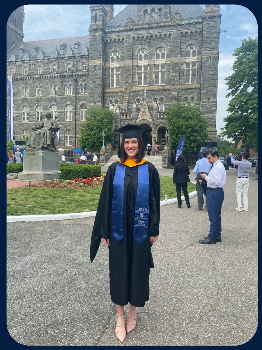 Gabriella stands in a black graduation robe and a bright blue stole. Healy Hall and Healy Lawn are behind her.