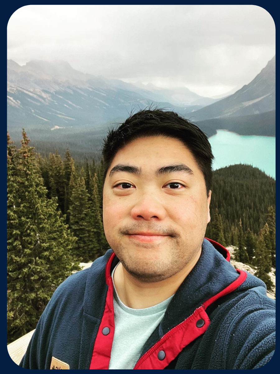 Matt smiles into the camera with a forest and a glacial lake behind him.
