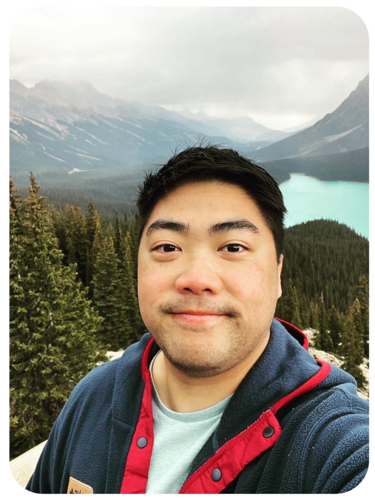 Matt smiles into the camera with a forest and a glacial lake behind him.