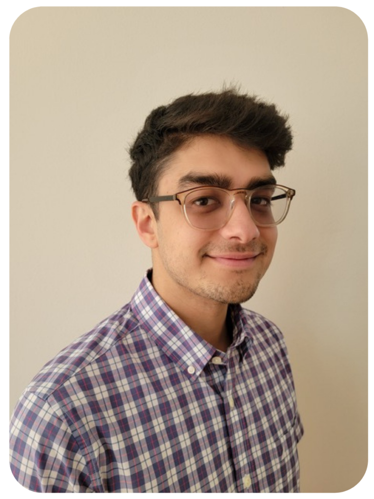 Adam smiles at the camera in front of a beige wall. He is wearing glasses and a plaid shirt.