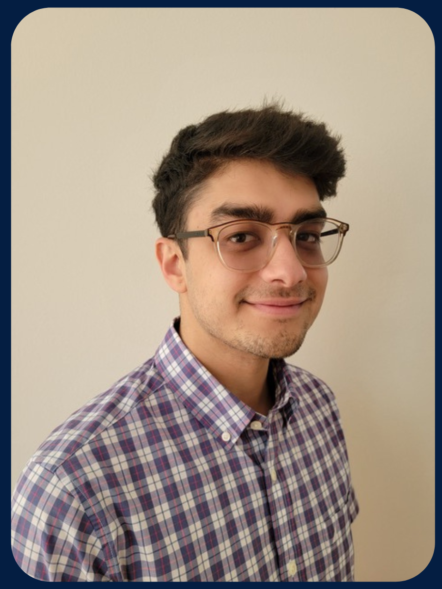 Adam smiles at the camera in front of a beige wall. He is wearing glasses and a plaid shirt.