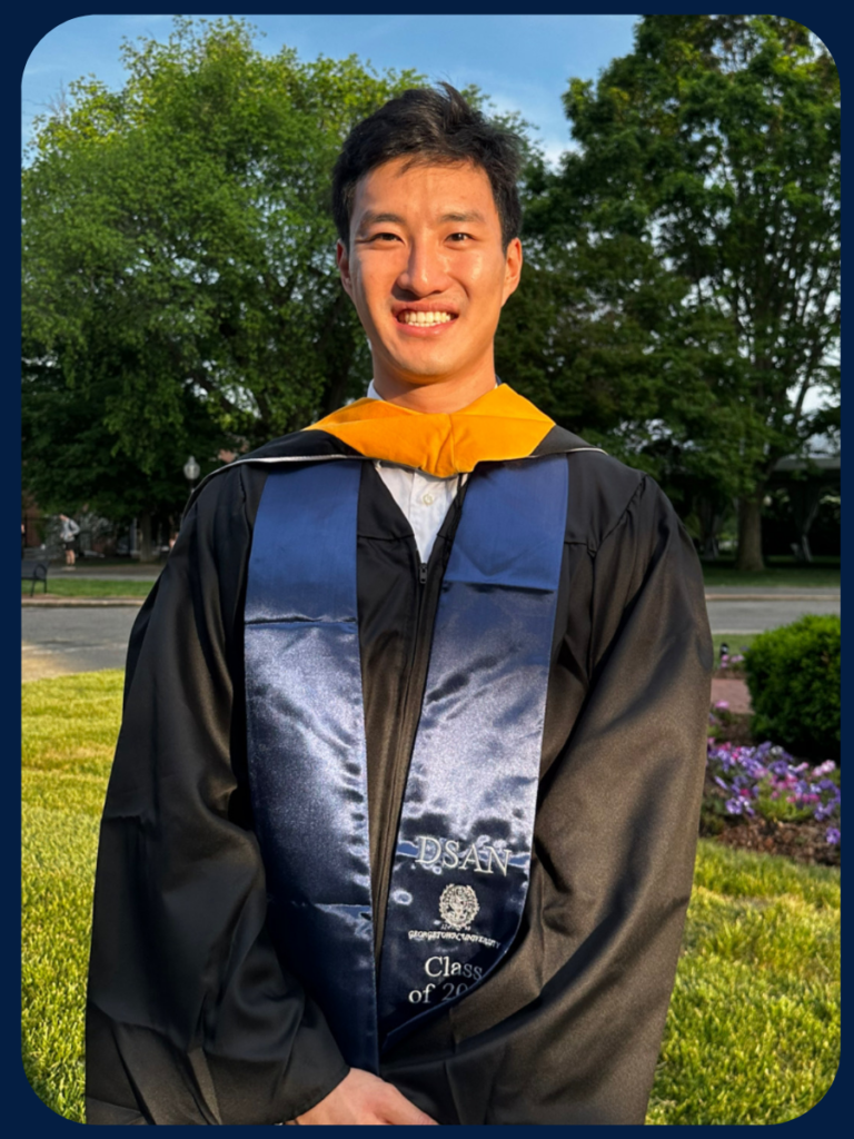 Brian Li is standing facing the camera smiling and wearing his black graduation gown with a yellow hood and blue stole.