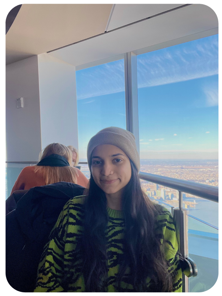 Anam sits in front of a window inside a high rise with the sky behind her. She has dark hair and his wearing a cream colored beanie and a green and black graphic-patterned sweater.