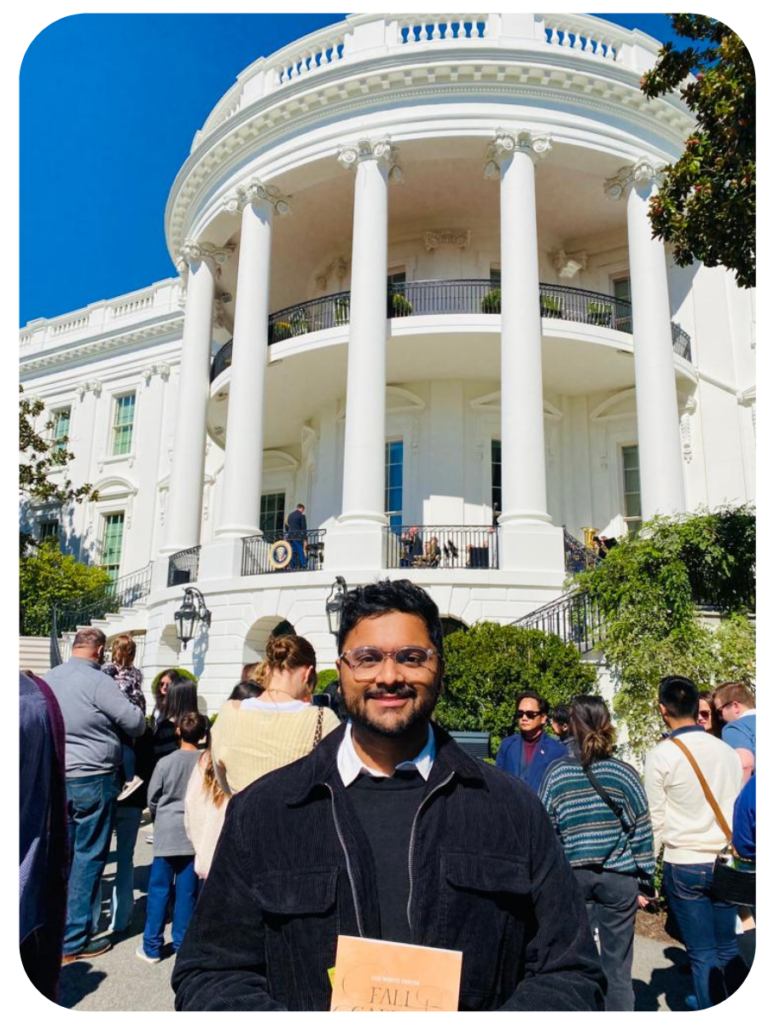 Swami Venkat stands on the South Lawn in front of the White House wearing a dark sweater with a white collared short underneath. People are milling around behind him. 