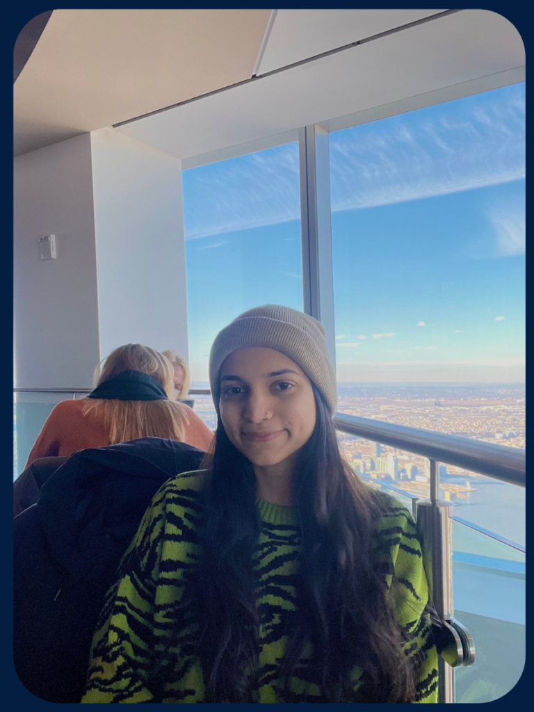 Anam sits in front of a window inside a high rise with the sky behind her. She has dark hair and his wearing a cream colored beanie and a green and black graphic-patterned sweater.