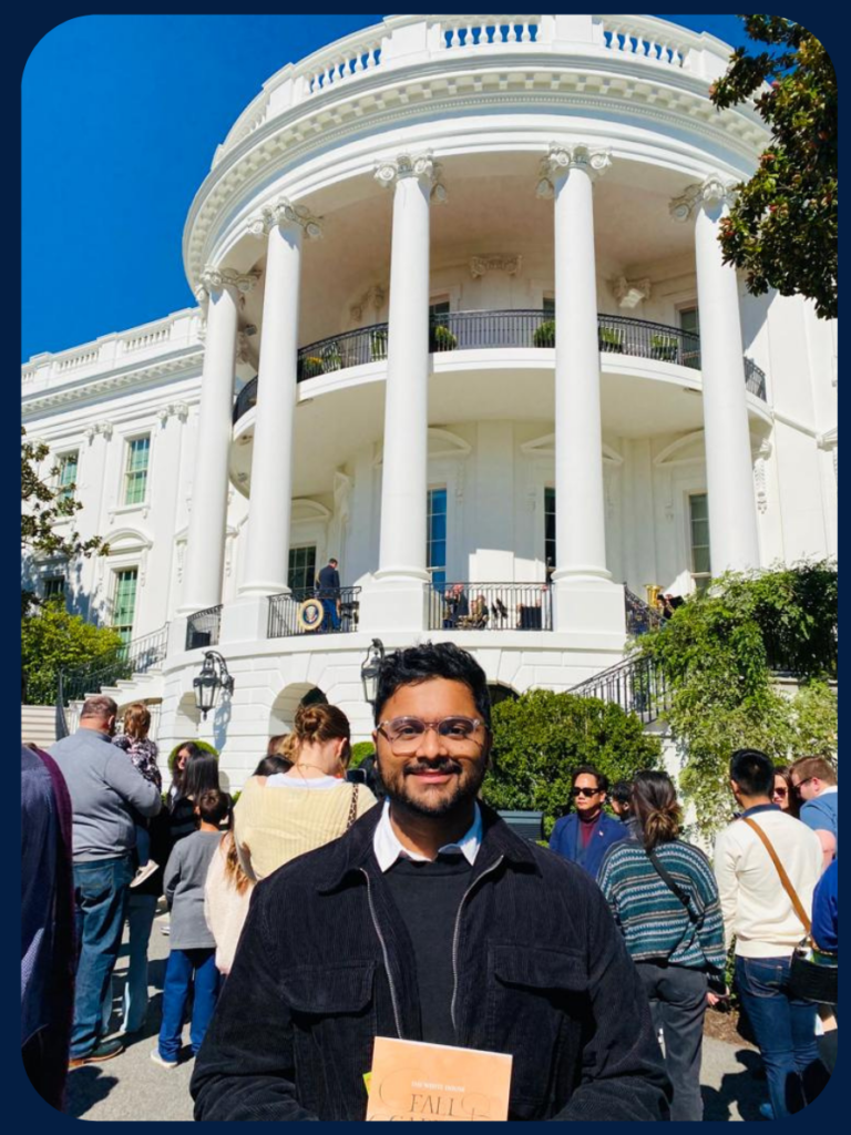Swami Venkat stands on the South Lawn in front of the White House wearing a dark sweater with a white collared short underneath. People are milling around behind him.