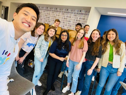 A large group of students smile facing the camera, standing in a large group. Brian Li is in the top left of the image holding the camera for a selfie.