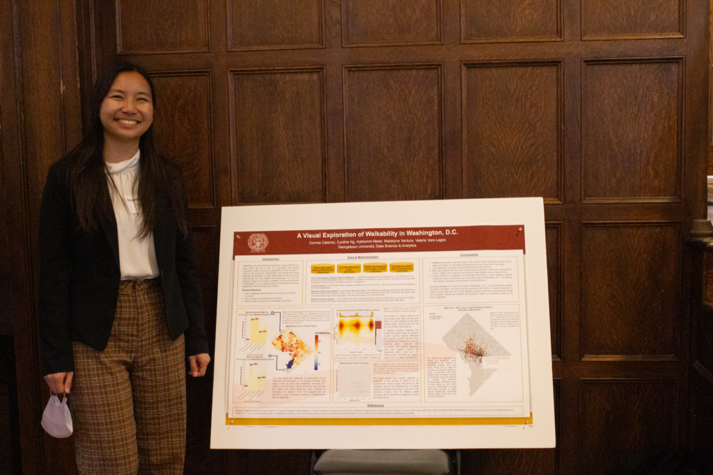 Cynthia stands to the left of her poster, in front of a wood-paneled wall.