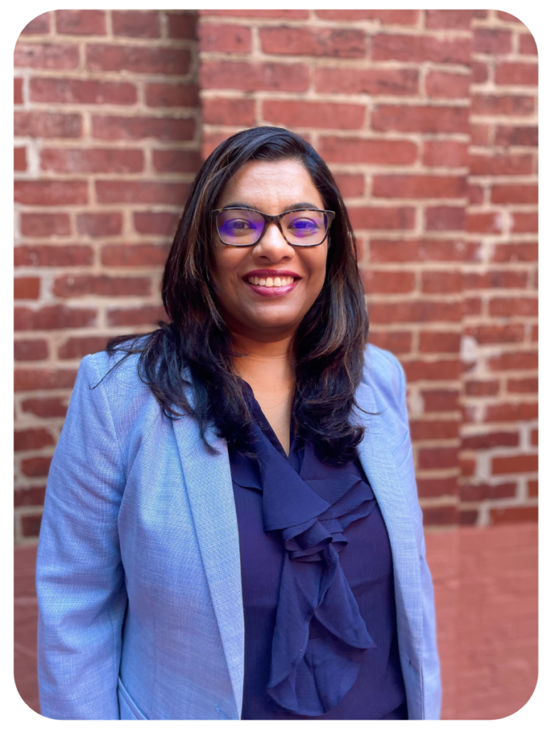 Purna stands smiling in front of a brick wall. She is wearing a navy blue blouse and light blue blazer.