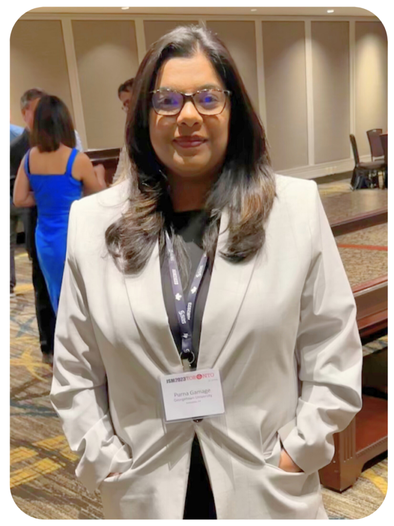 Purna stands in a large room at a conference, smiling at the camera. She is wearing a taupe blazer and black shirt.