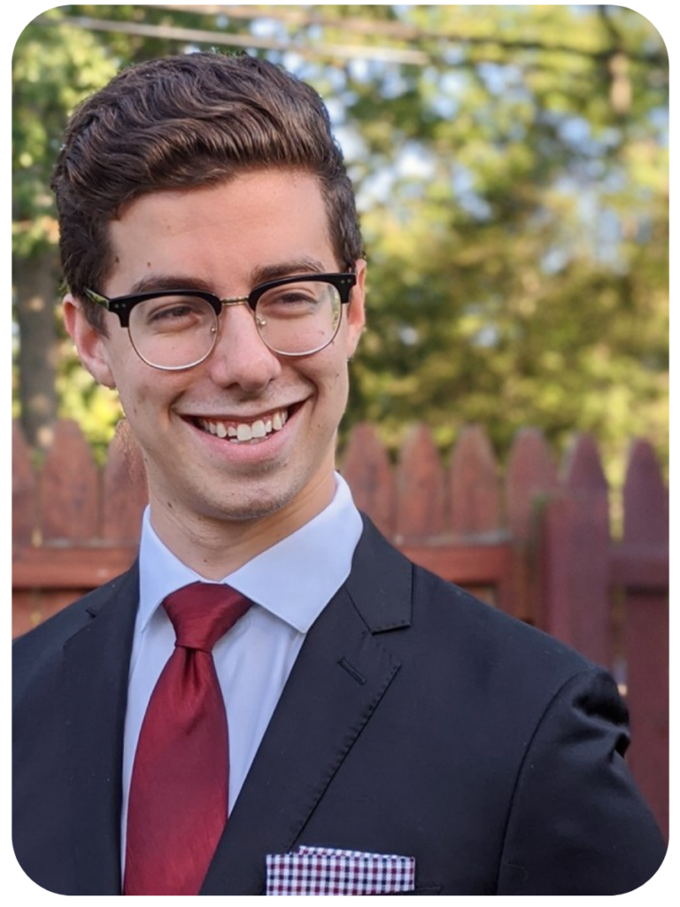 Zev, a brown-haired man, smiles in front of a wooden fence. He is wearing a navy blue suit, light blue shirt, dark red tie, and a patterned pocket square.