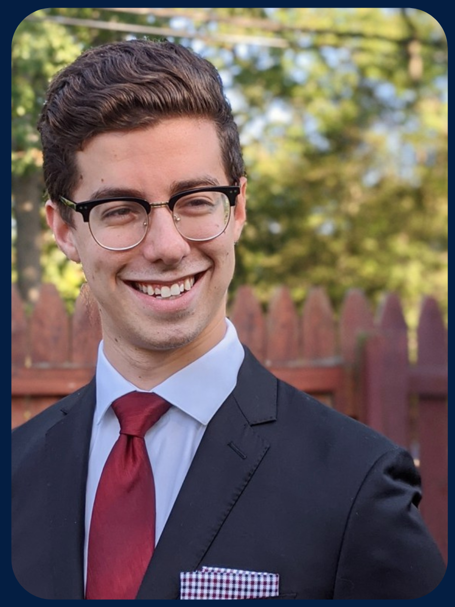 A brown-haired Zev smiles in front of a wooden fence. He is wearing a navy blue suit, light blue shirt, dark red tie, and a patterned pocket square.
