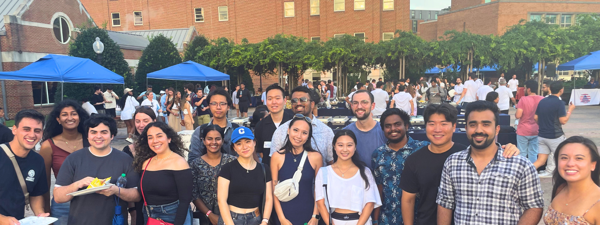 Students stand smiling facing the camera standing outdoors under a sunny sky. Food tents and other students stand in groups behind them.