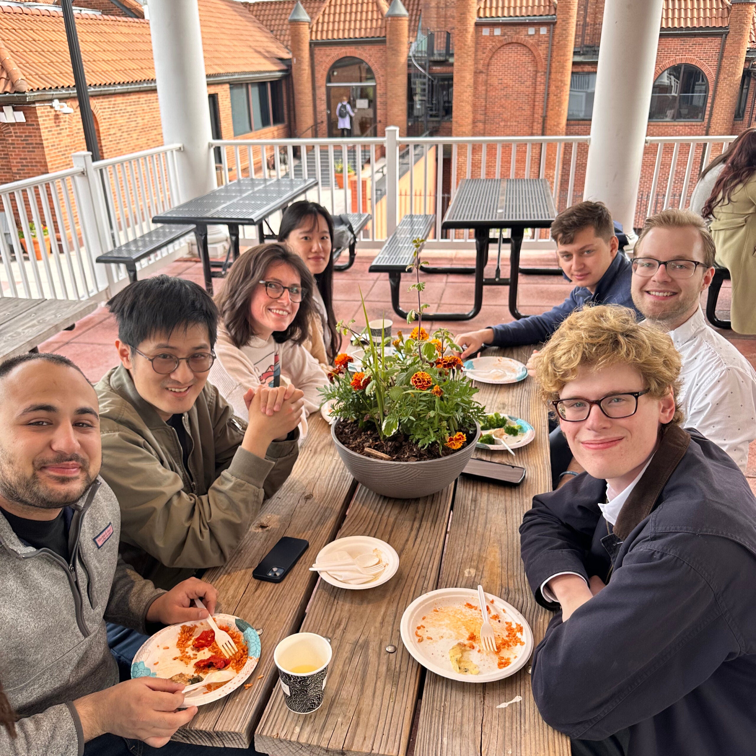 7 students sit on either side of a picnic table eating are facing the camera, smiling.