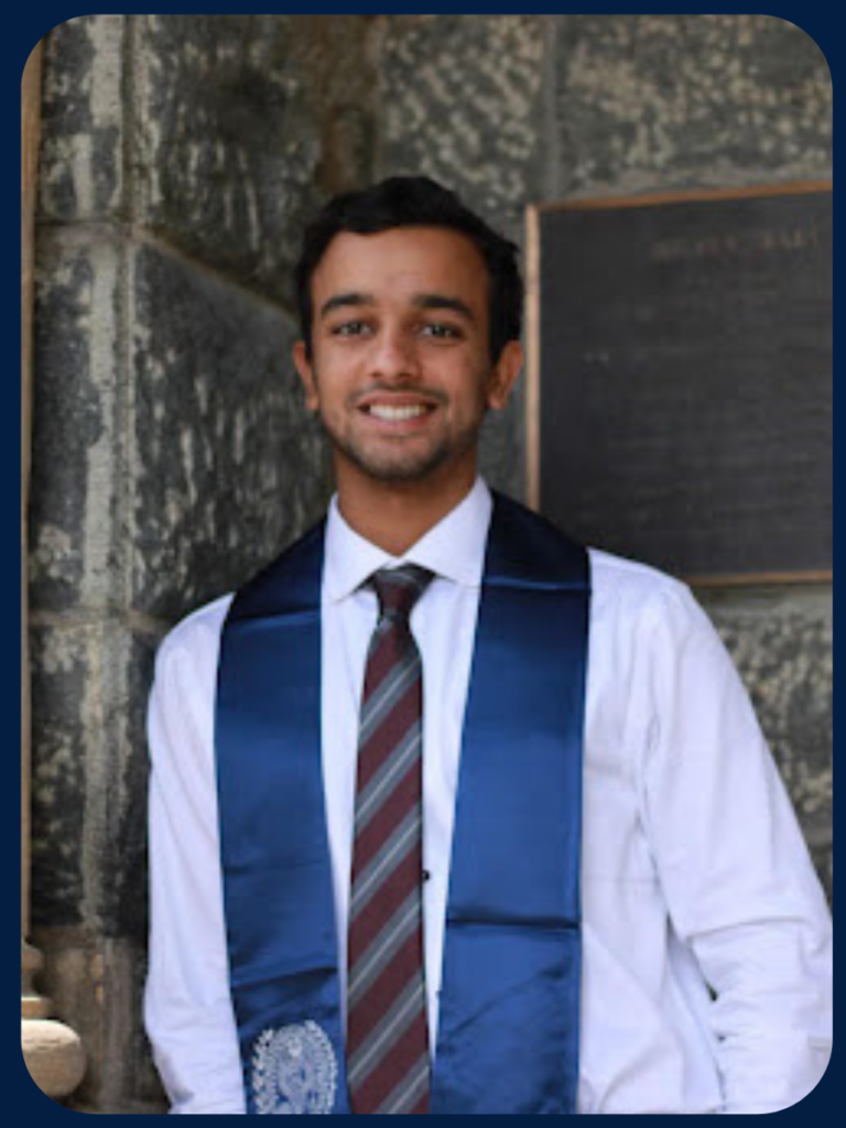 Raunak stands next to a stone pillar wearing a white button-down shirt, striped tie, and blue Georgetown graduation stole.