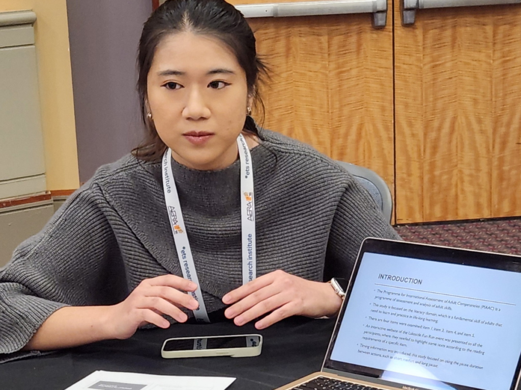 Yiming sits at a table with her open laptop showing her research work. She is wearing a gray sweater.