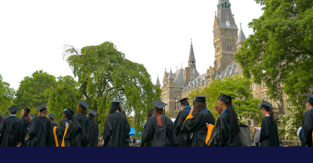 Students in cap and gown walk in the foreground with trees and Healy Hall in the background.